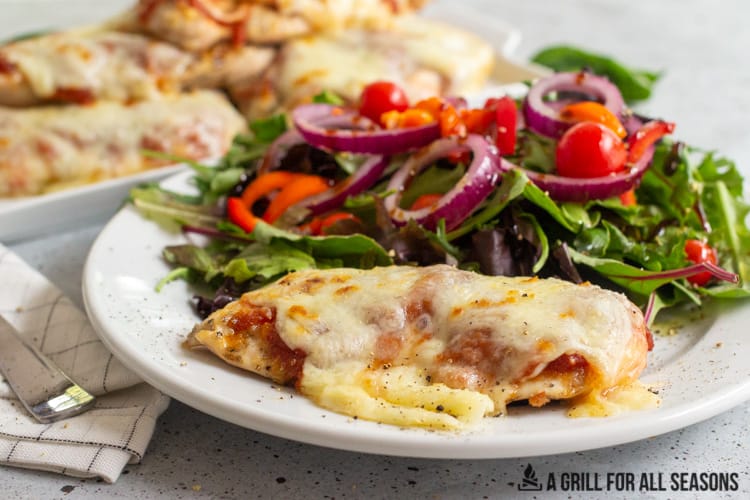 tray of grilled chicken parm and dish with piece of chicken parm accompanied by a garden salad.