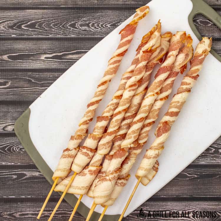 Raw bacon on a stick. Wrapped slices of bacon around bamboo skewers on a cutting board.