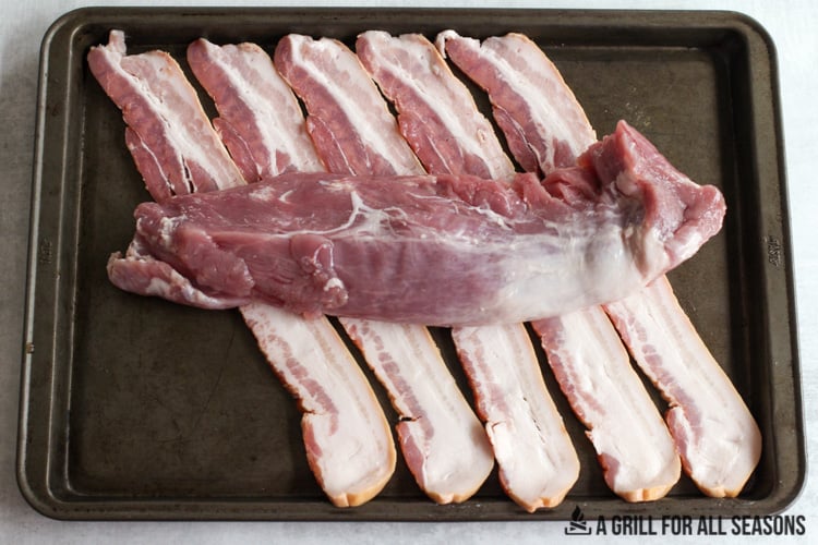 Pork tenderloin being wrapped in bacon on cooking sheet.