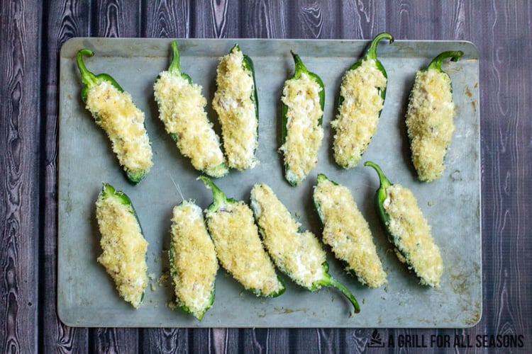 Jalapenos filled with cream cheese mixture, topped with bread crumbs on baking sheet.