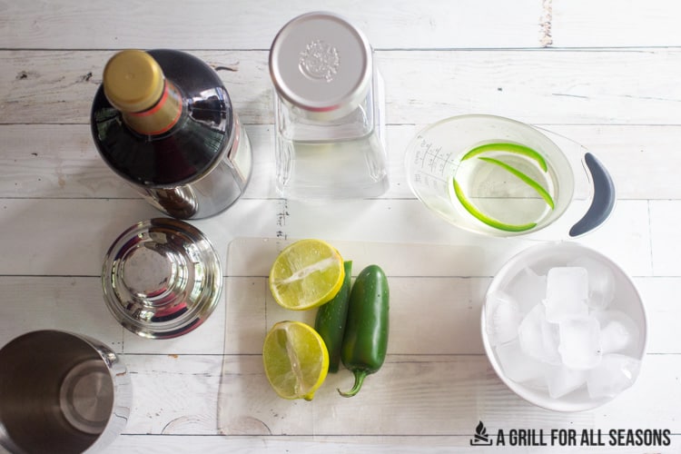Assembled ingredients of Triple Sec, Tequila, Cocktail Shaker, limes, jalapenos, simple syrup and ice cubes in a bowl.