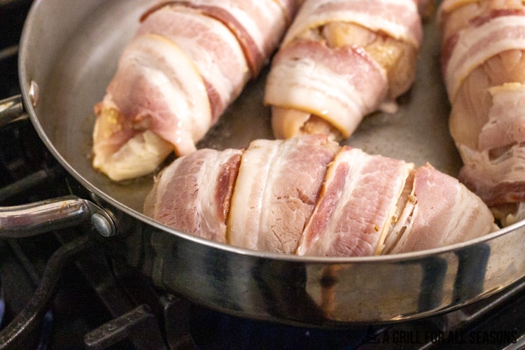 Bacon wrapped stuffed chicken being seared in pan on stove.