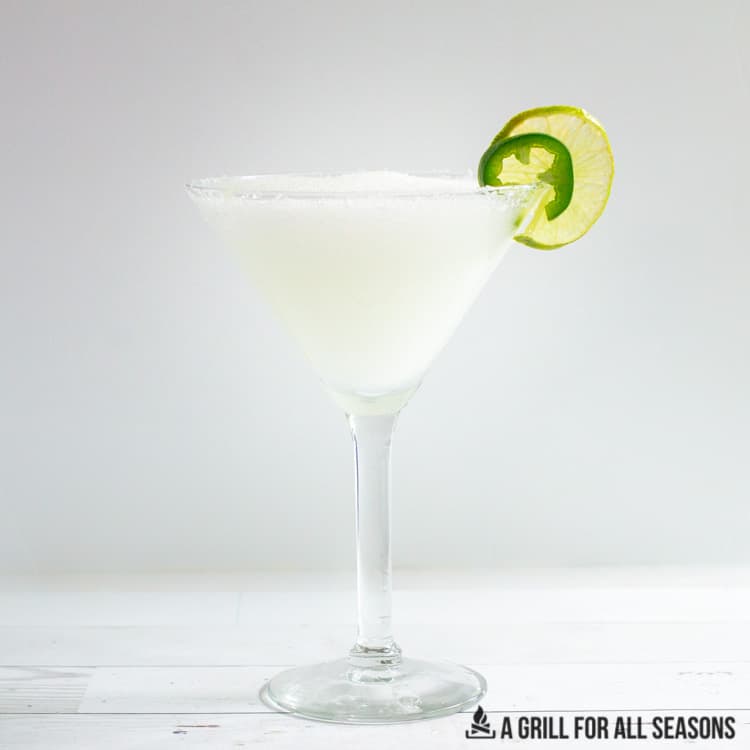 Frozen margarita being poured into cocktail glass with slices of lime and jalapeno as garnishes.