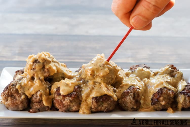 Venison meatballs on white serving platter with bourbon cream sauce poured over them. One of them being speared with a toothpick.