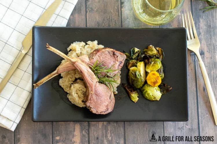 Two lamb chops sitting on bed of quinoa on plate with brussel sprouts and sprig of rosemary. Glass of white wine and utensils in back ground.