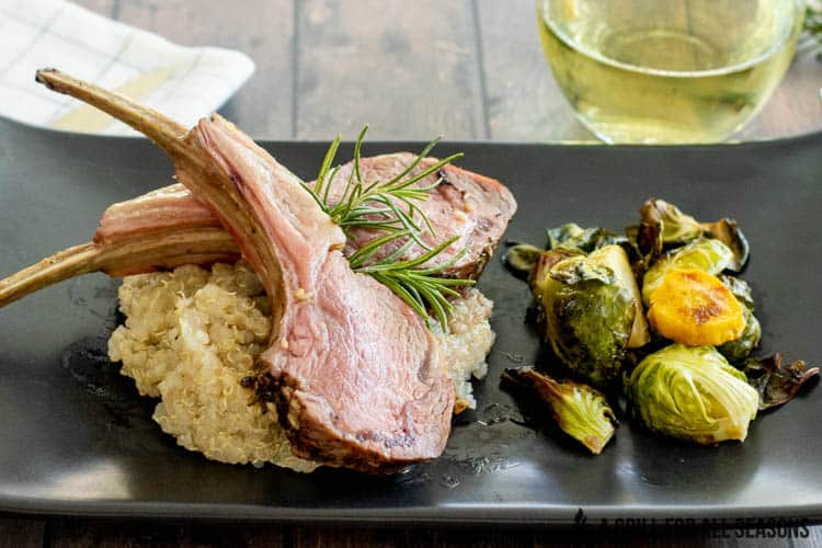 Two lamb chops sitting on bed of quinoa on plate with brussel sprouts and sprig of rosemary. Glass of white wine in background.