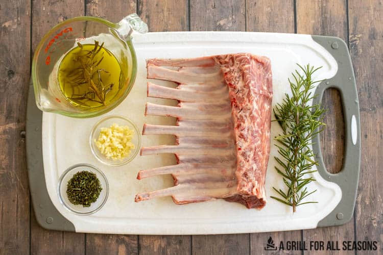 Olive oil, fresh rosemary, garlic, and rack of lamb sitting on cutting board.