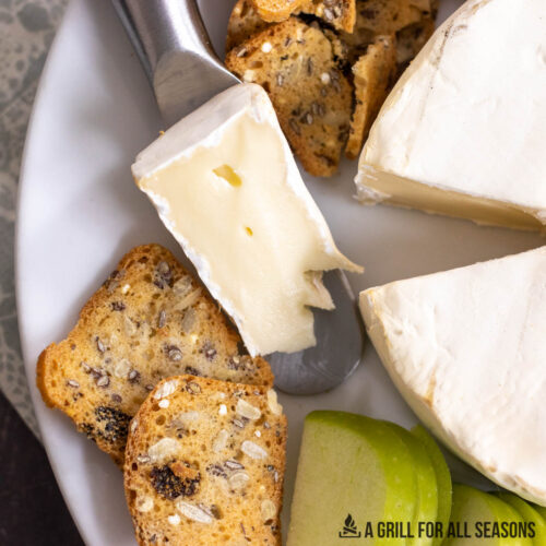 smoked brie cheese wedge on serving knife next to wheel of brie and crackers