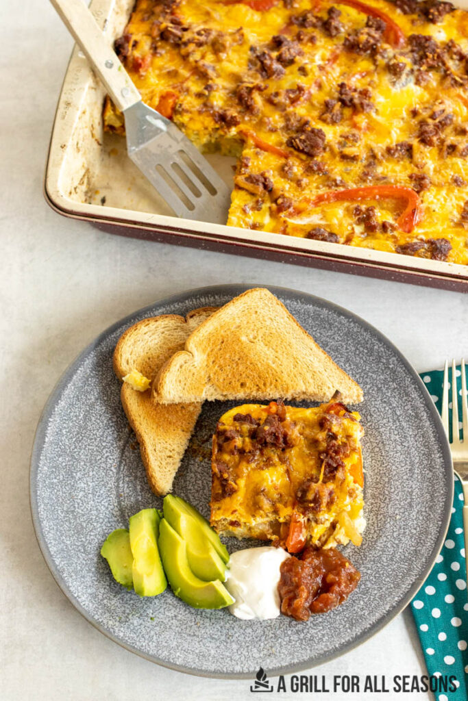chorizo casserole served on plate with toast, avocado, sour cream, and salsa with baking pan behind