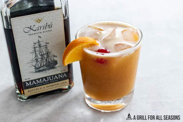 mamajuna cocktail garnished with orange and cherry alongside bottle fo mamjuana rum