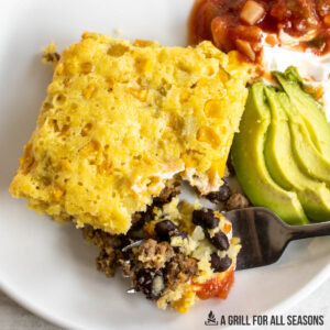 mexican cornbread casserole recipe on plate with salsa, sour cream, and avocado with a bite on a fork