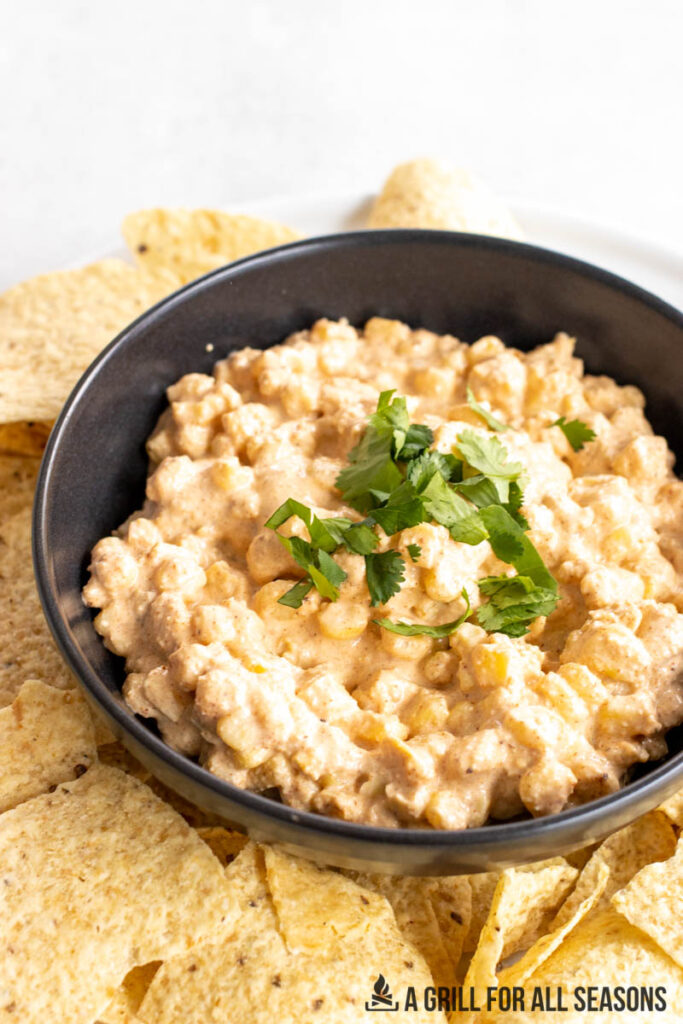 vertical close up of elote dip recipe in a black bowl surrounded by tortilla chips