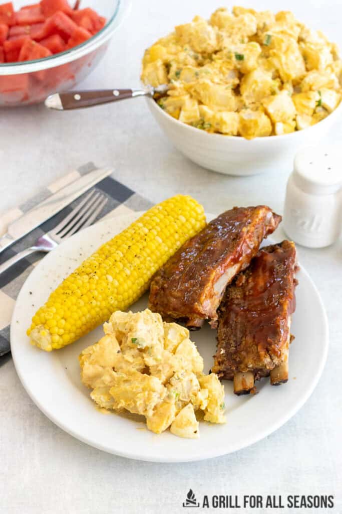 southern potato salad on plate with bbq ribs and corn on the cob with bowls behind