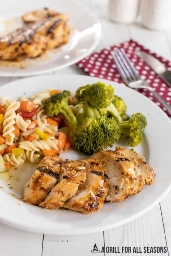 slices of honey mustard grilled chicken breast on dinner plate with pasta salad and broccoli and serving platter behind