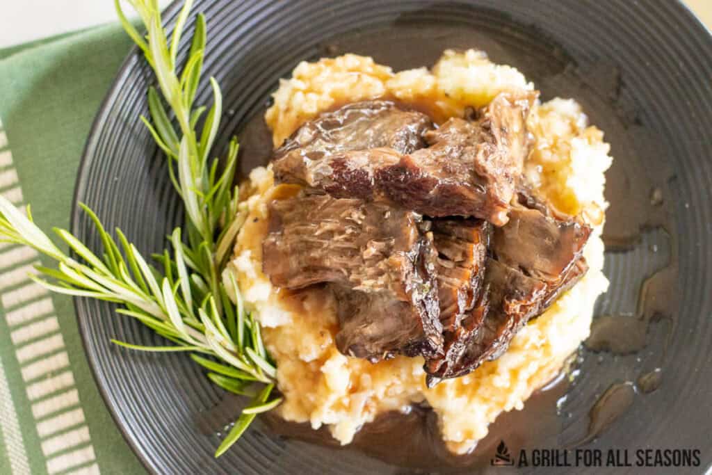 braised chuck roast with gravy served on mashed potatoes on dinner plate with rosemary garnish