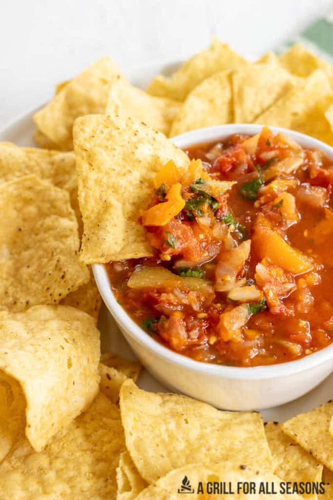 vertical shot of bowl of smoked salsa with chip dipped in it in the middle of a platter of tortilla chips